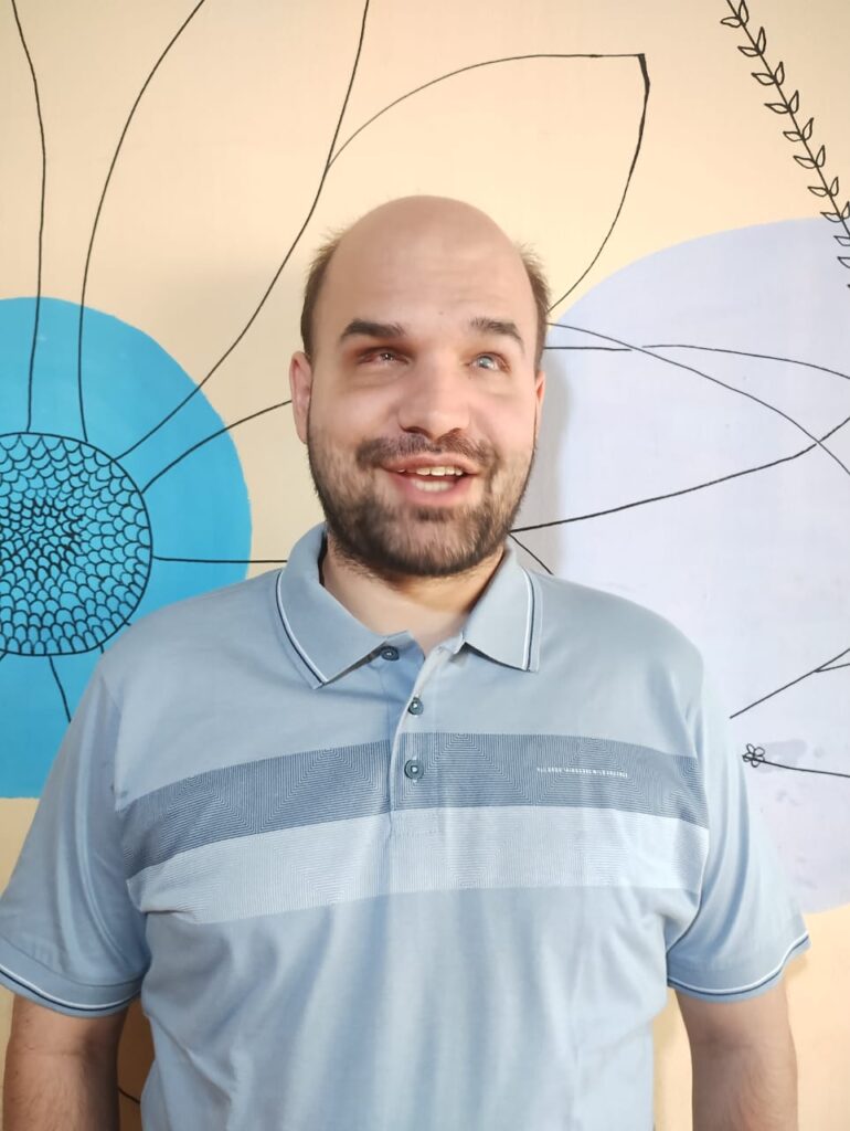 Jean aparece sorrindo de forma aberta, com expressão simpática e animada. Ele usa uma camisa polo azul-clara com listras horizontais em tons de azul no peito.